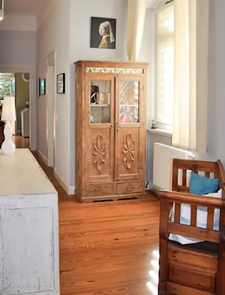 Wohnzimmer mit Holzschrank, Kinderbett, Holzboden und Wandbild unter hellem Fenster.