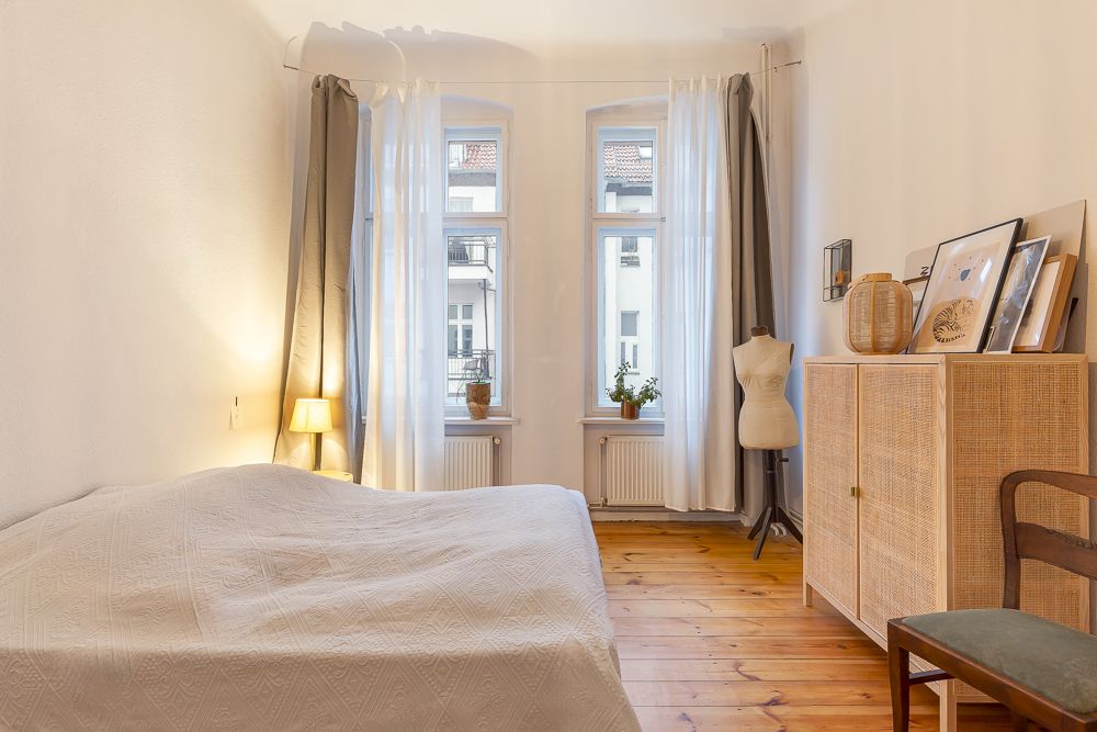 Helles Schlafzimmer mit Bett, Holzboden, großer Kommode, zwei Fenstern und Vorhängen.