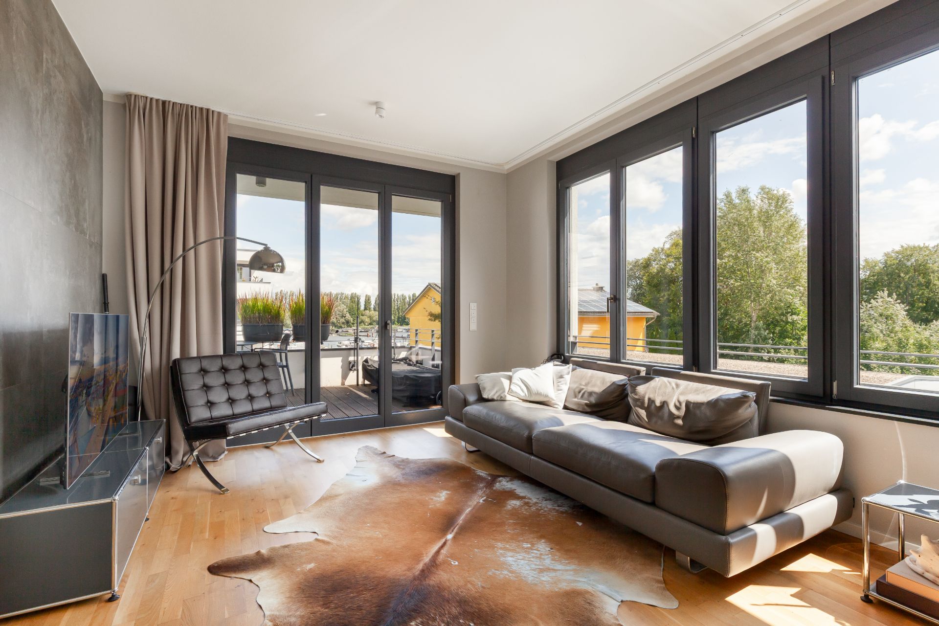 Modernes Wohnzimmer mit großer Fensterfront, grauem Sofa und Kuhfell-Teppich auf Holzfußboden.