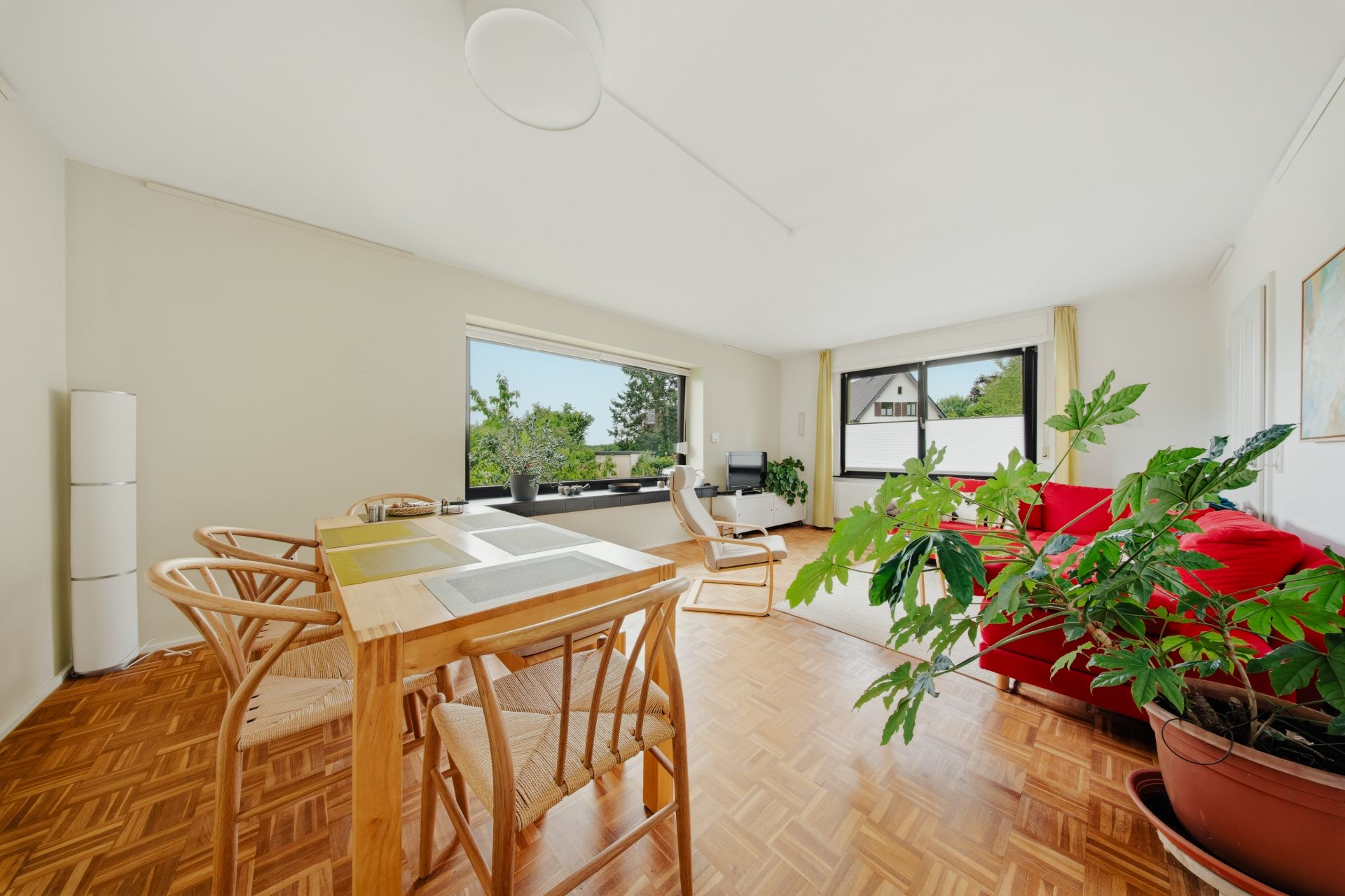 Helles Wohnzimmer mit roten Sofa, Esstisch, Stühlen, großen Fenstern und Zimmerpflanze.