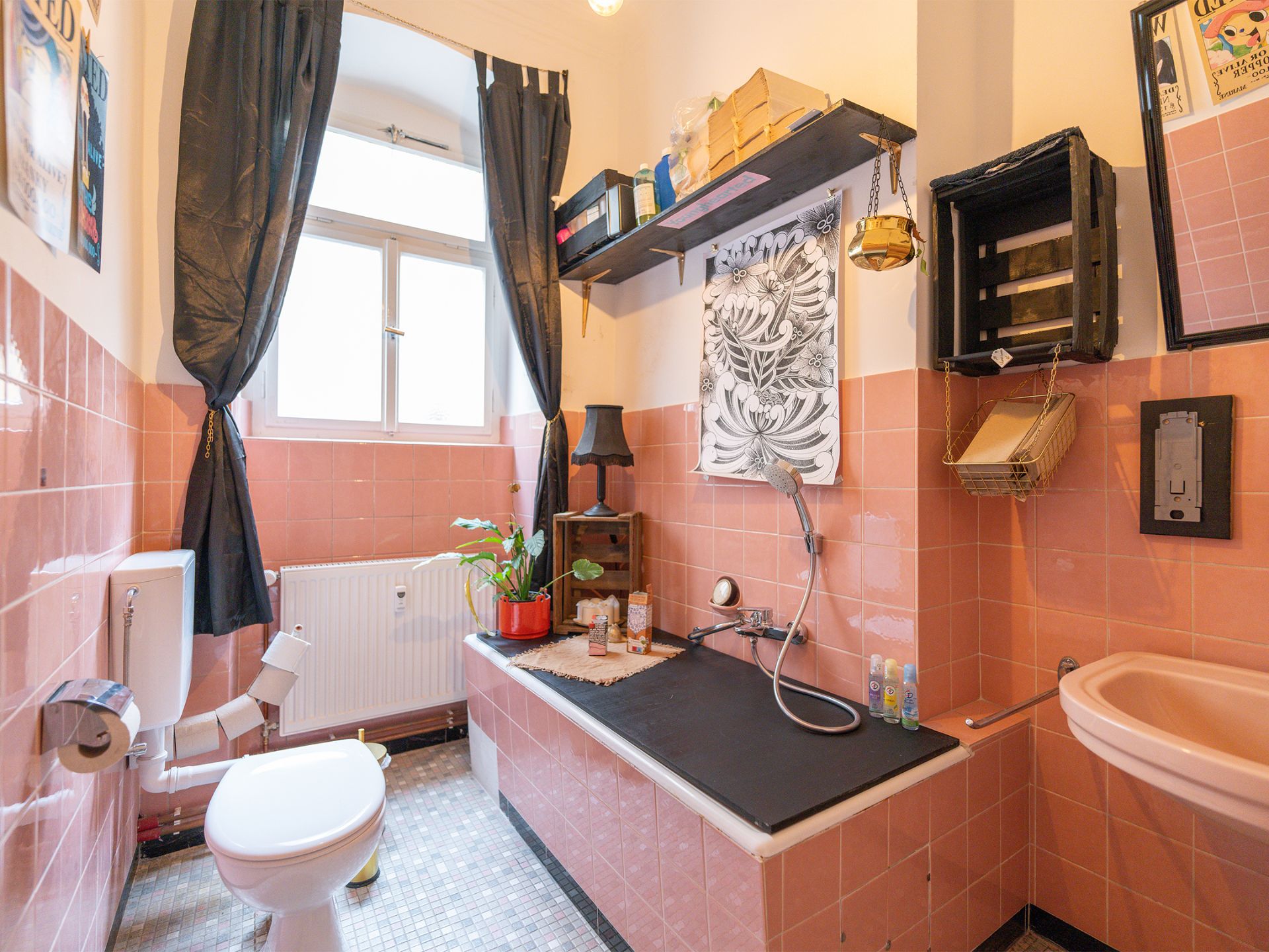 Kleines Badezimmer mit rosa Fliesen, Badewanne, Toilette, Waschbecken und Fenster mit schwarzen Vorhängen.