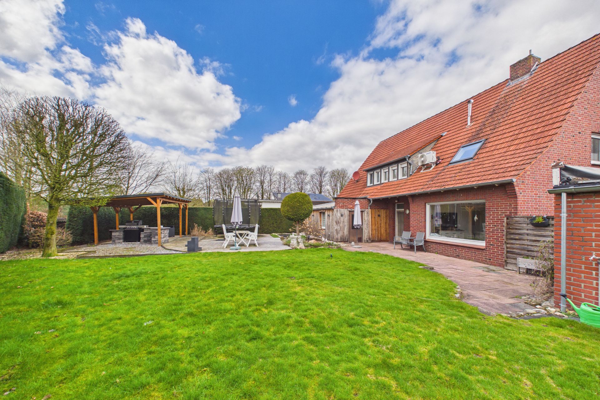 Großer Garten mit Rasen, Terrasse, Gartenmöbeln und rotem Wohnhaus mit schrägem Dach.