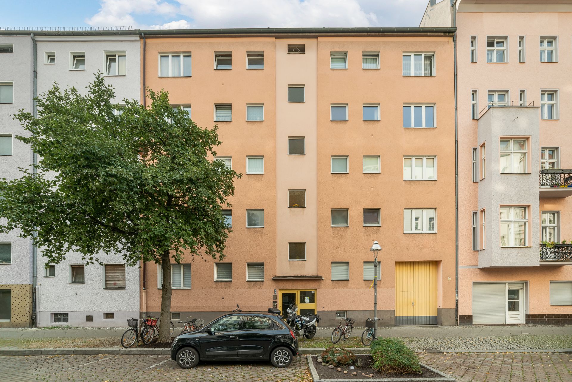 Vorderansicht eines mehrstöckigen Wohnhauses mit oranger Fassade, Baum, geparktem Auto und Fahrrädern davor.