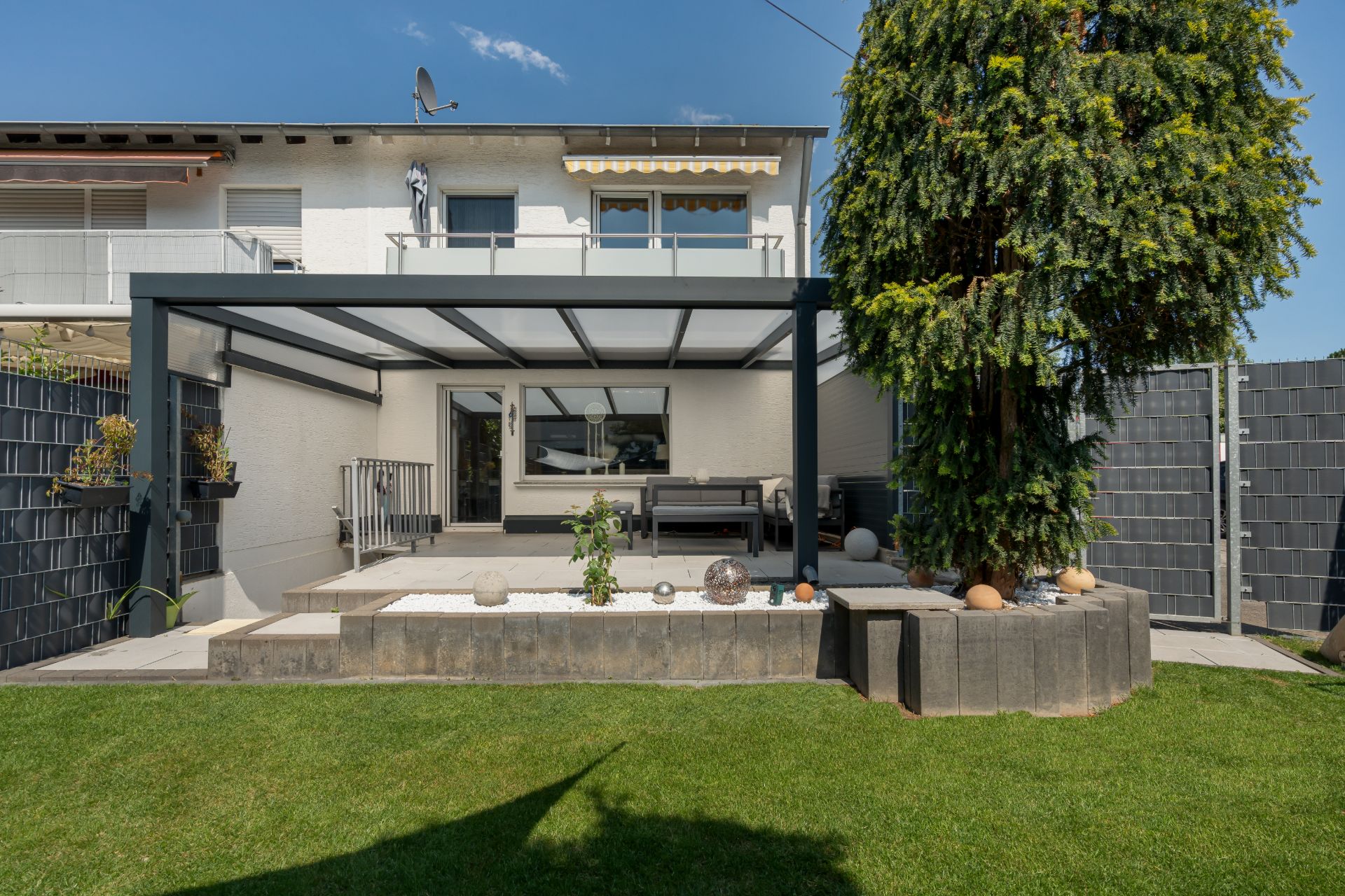 Moderne Terrasse mit Glasdach, Sitzecke, kleinem Garten und großem Baum im Garten eines Wohnhauses.