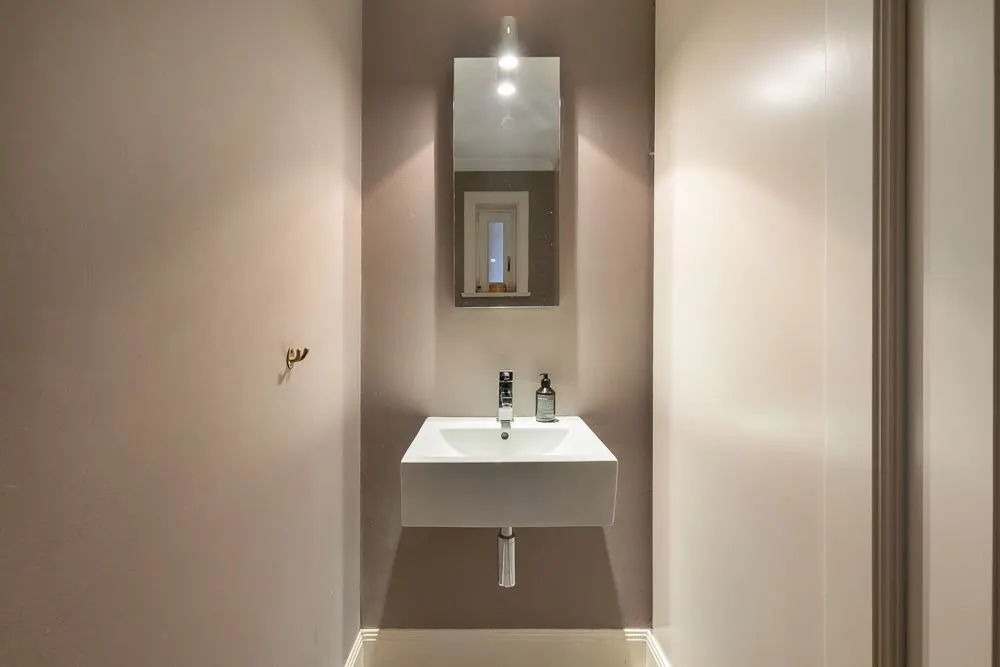 Schmales Badezimmer mit Waschbecken, Spiegel und Seifenspender, beleuchtet von einer Deckenlampe.