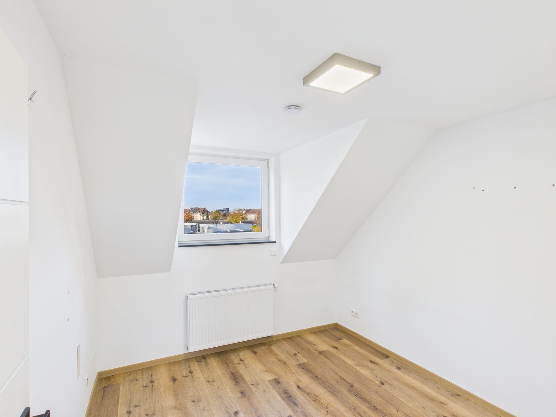 Helles, leeres Zimmer mit Dachschräge, Fenster, Heizkörper und Holzfußboden.