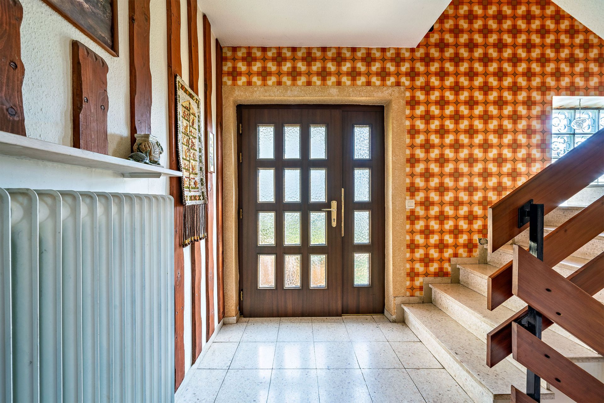 Hausflur mit brauner Eingangstür, gemusterter Tapete und Treppe mit Holzgeländer.