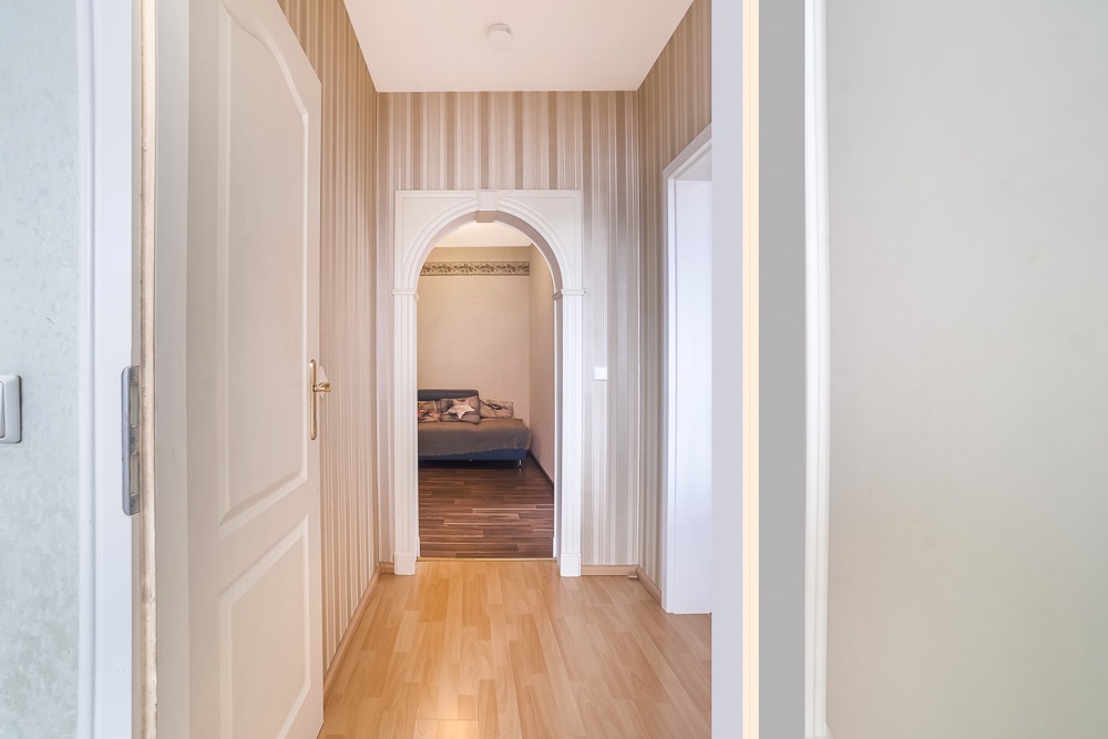 Flur mit Holzfußboden, offener Tür und Blick auf ein Schlafzimmer durch einen Torbogen.