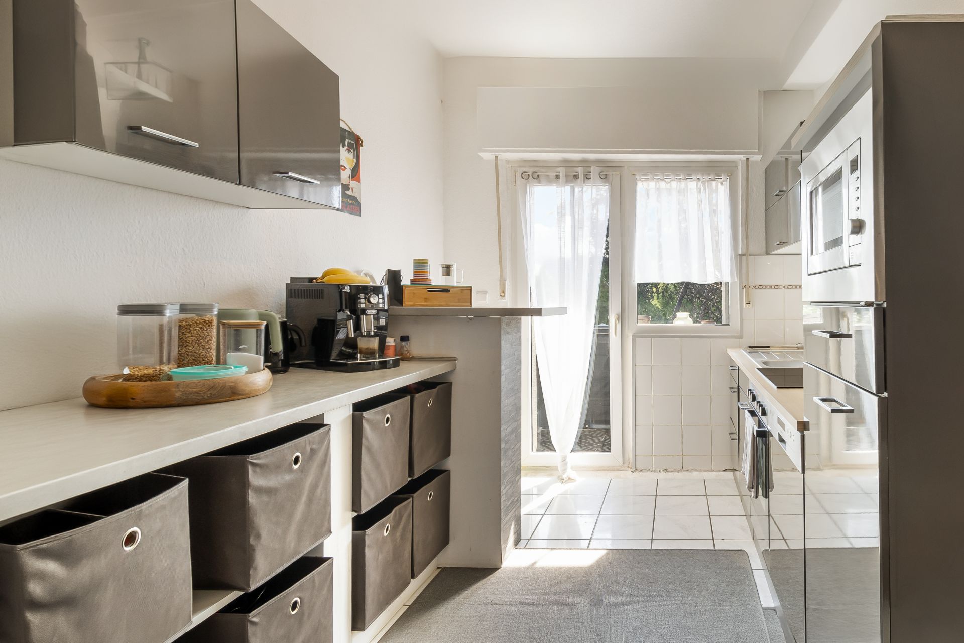 Helle Küche mit grauen Schränken, Aufbewahrungsboxen, Kaffeemaschine und Zugang zum Balkon.