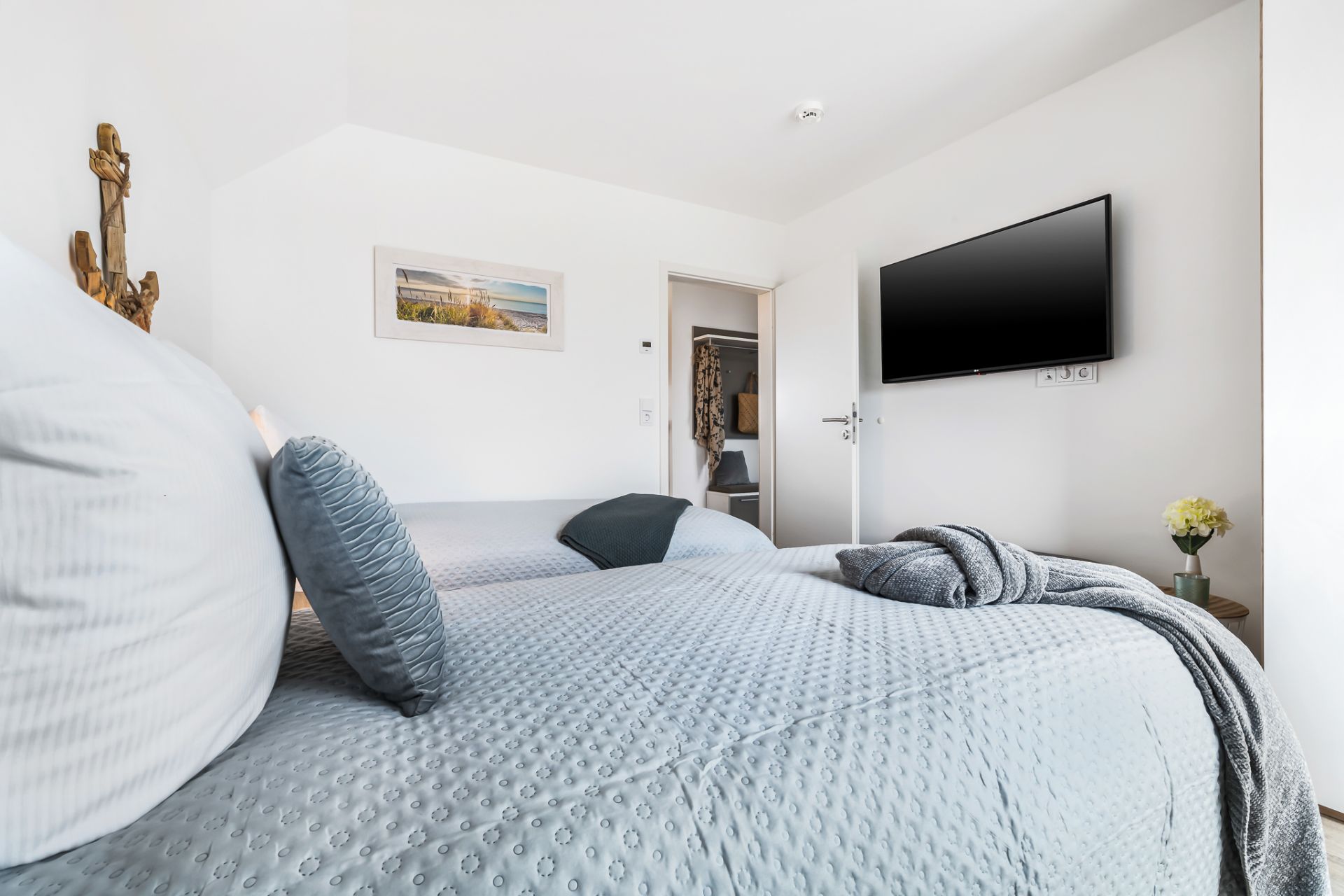 Helles Schlafzimmer mit Doppelbett, Wandbild, Fernseher und kleiner Vase mit Blume auf einem Nachttisch.