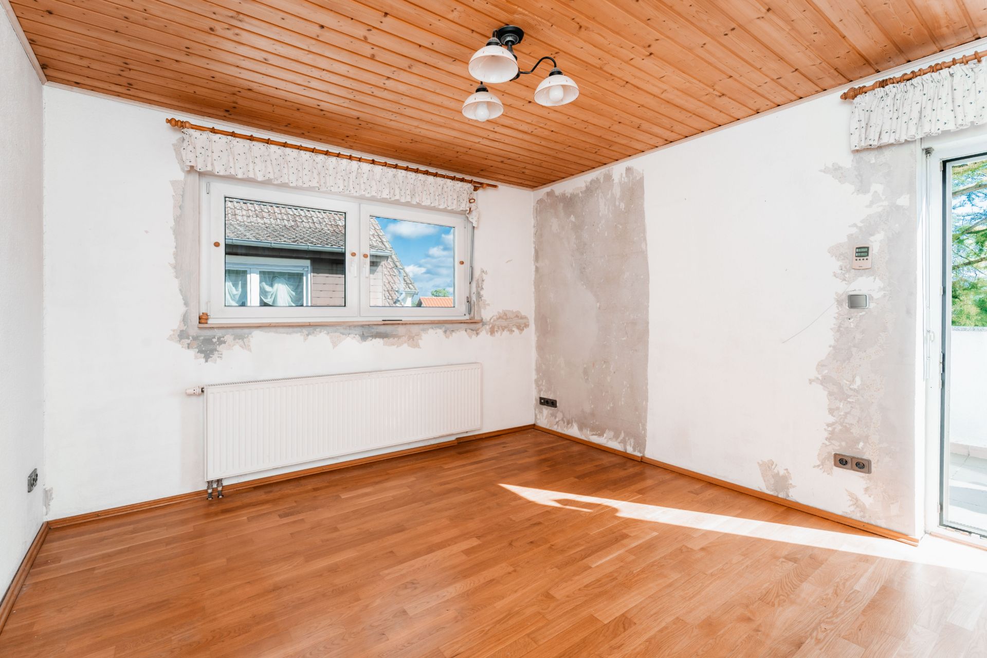 Leerer Raum mit Holzboden, Holzdecke, Fenster, Heizung und sichtbaren Wandschäden.