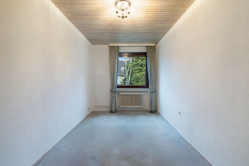 Leerer, schmaler Raum mit weiß gestrichenen Wänden, Deckenleuchte und Fenster mit Vorhängen.