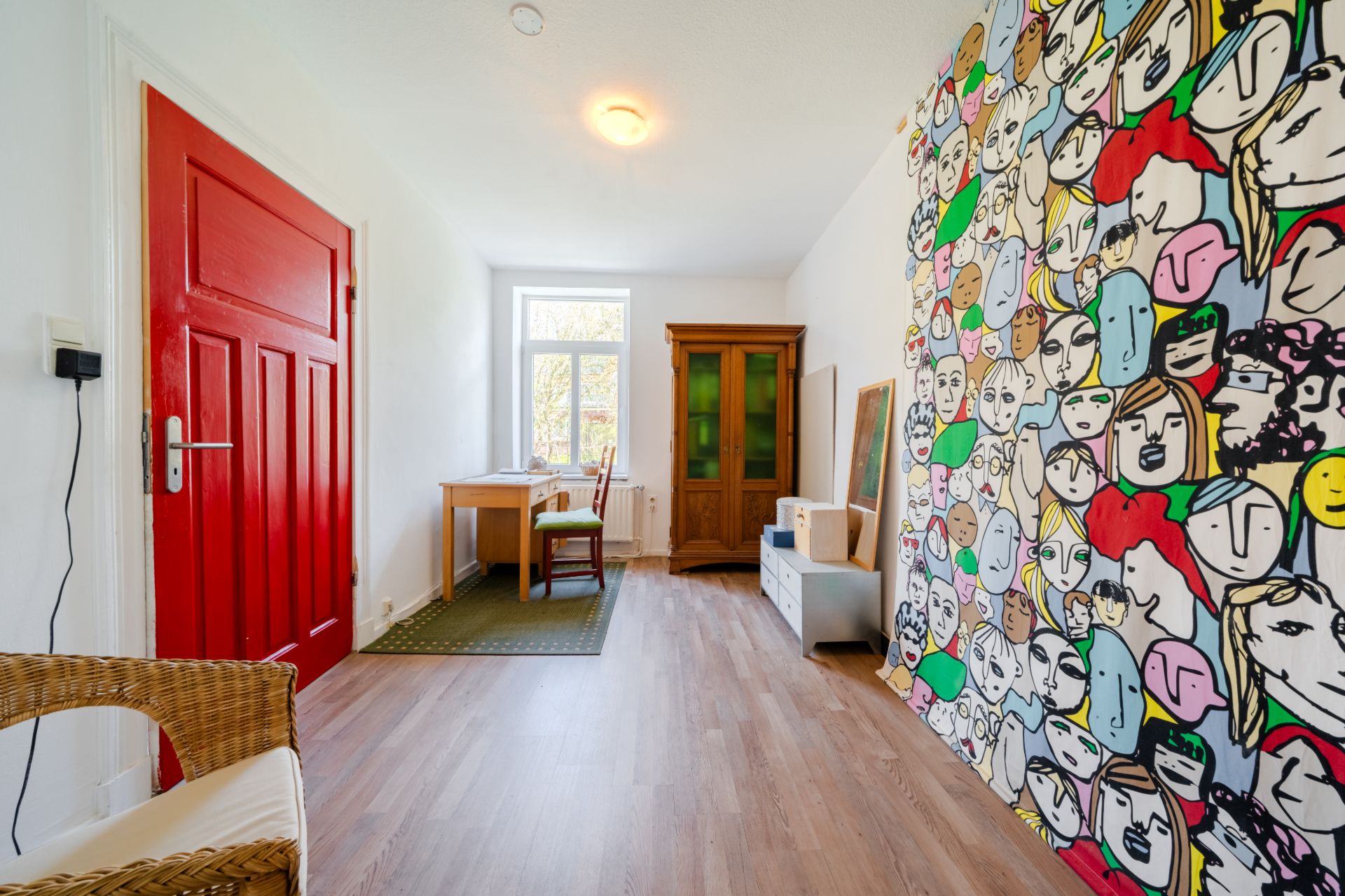 Helles Zimmer mit roter Tür, buntem Wandbild, Schreibtisch, Stuhl und Holzschrank.