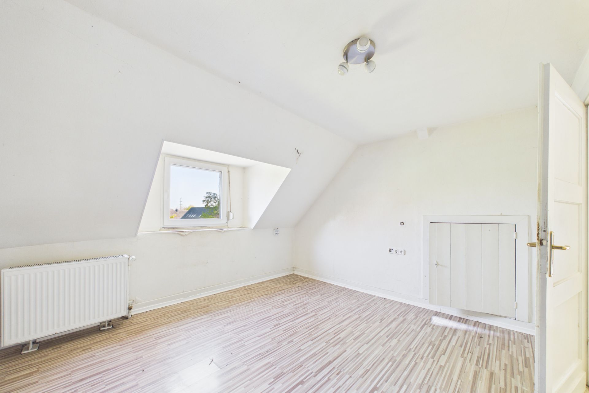 Helles, leeres Dachzimmer mit Fenster, Heizung und Lampe an der Decke.