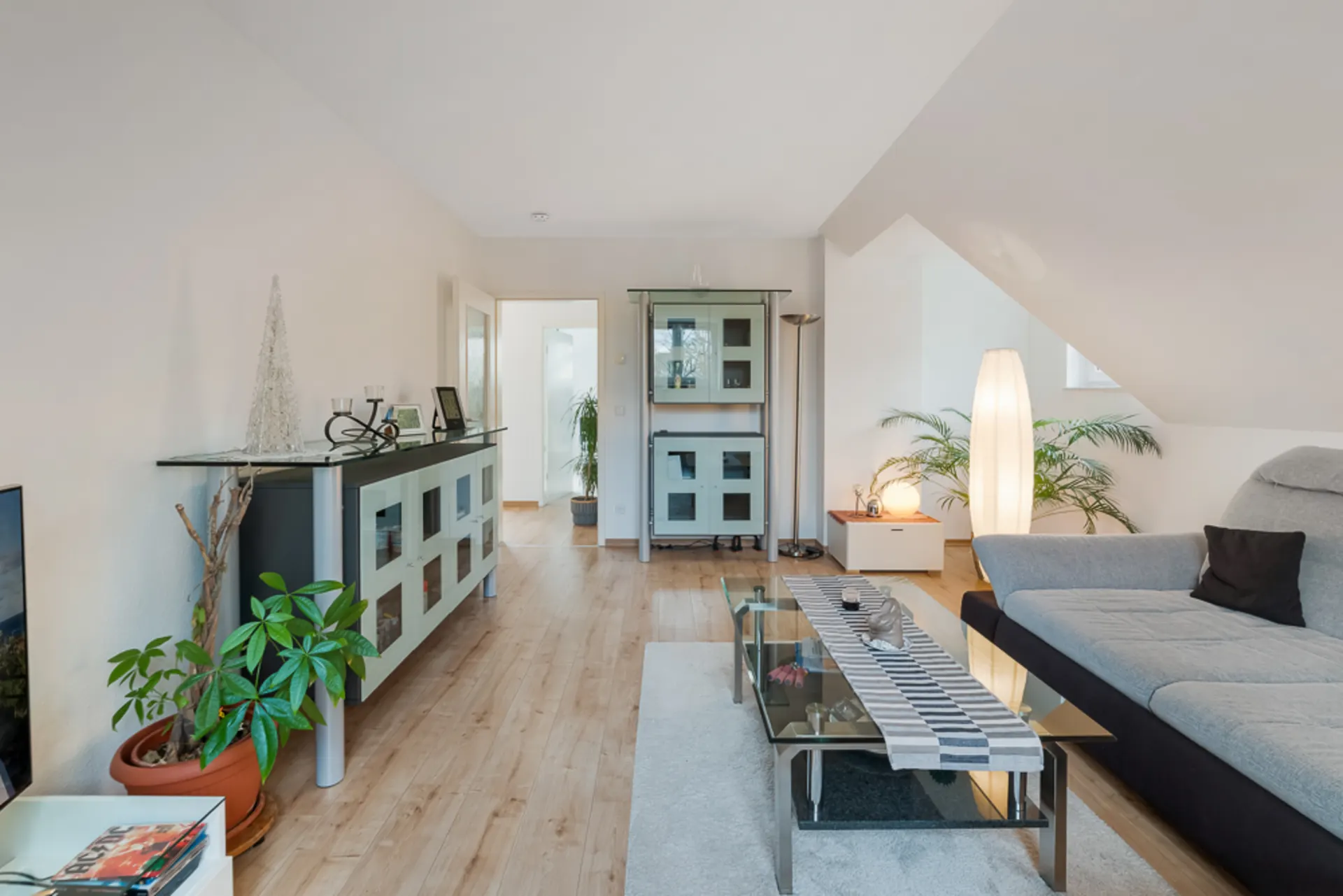 Helles Wohnzimmer mit grauem Sofa, Glastisch, Sideboard, Pflanzen und Stehlampe.