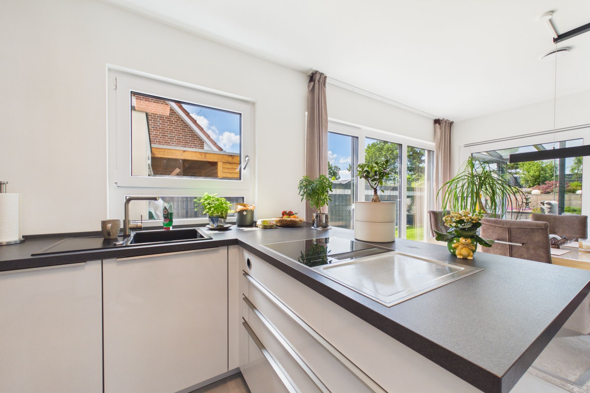 Moderne Küche mit großen Fenstern, Pflanzen und Essbereich im Hintergrund.