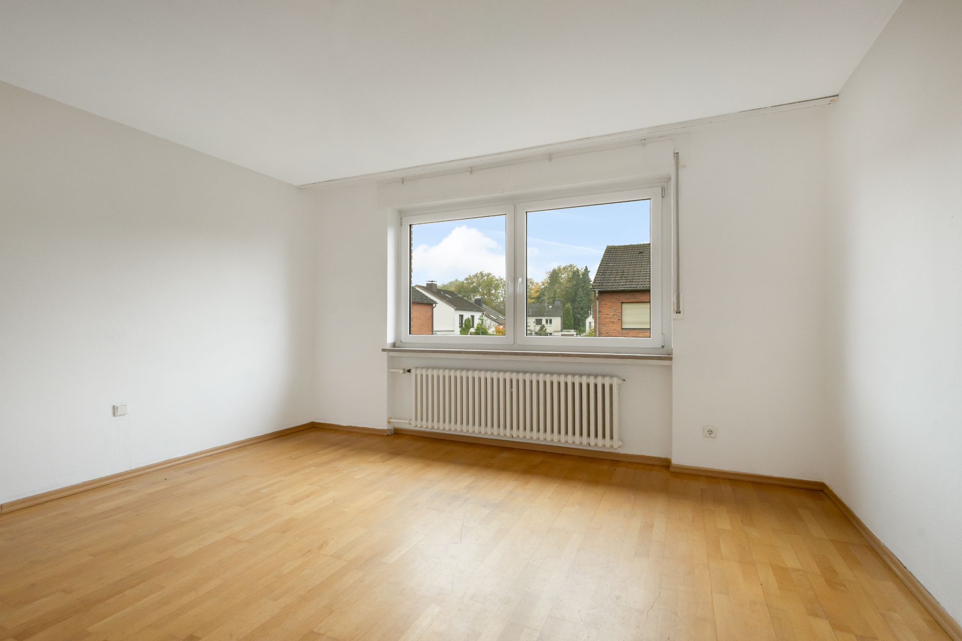 Leerer Raum mit Holzfußboden, weißer Wand und großem Fenster mit Blick nach draußen.