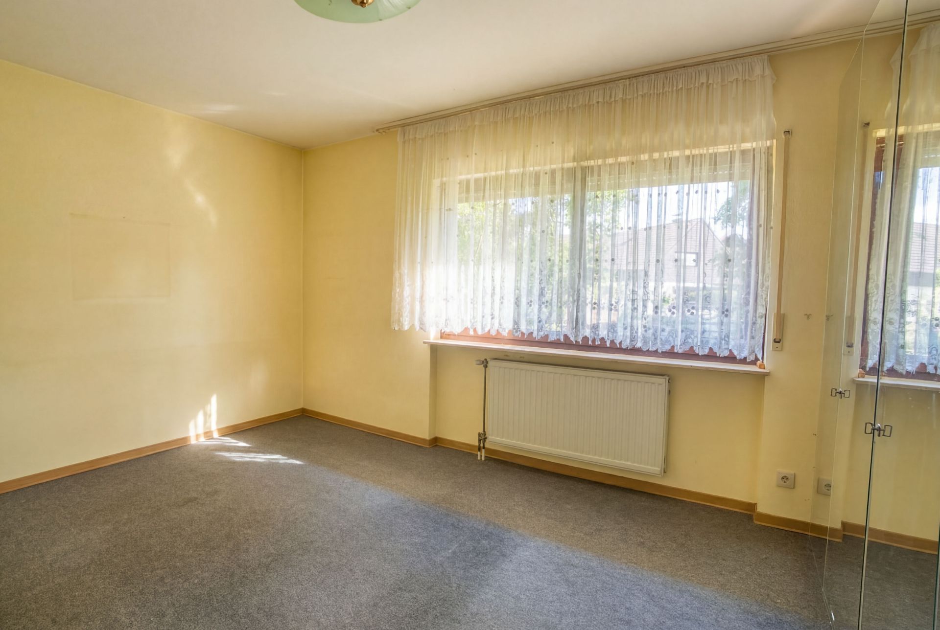 Leeres Zimmer mit gelber Wand, Teppichboden, großem Fenster mit Gardinen und Heizkörper.