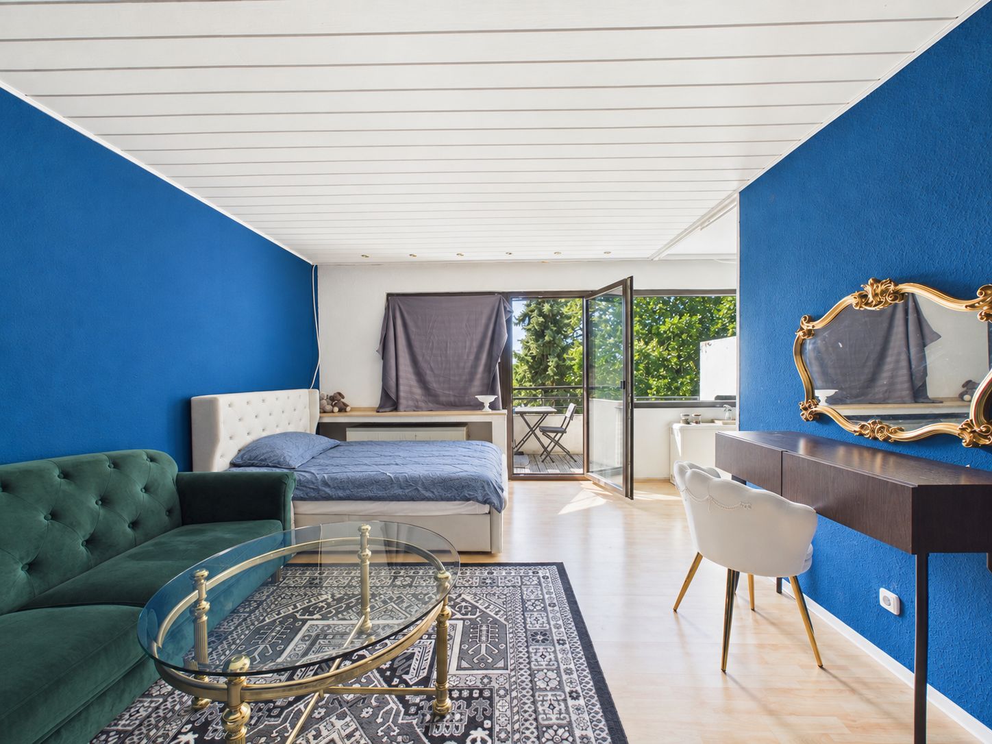 Wohnzimmer mit grünem Sofa, Bett, Glastisch, Schreibtisch und offenem Balkon, blaue Wand.