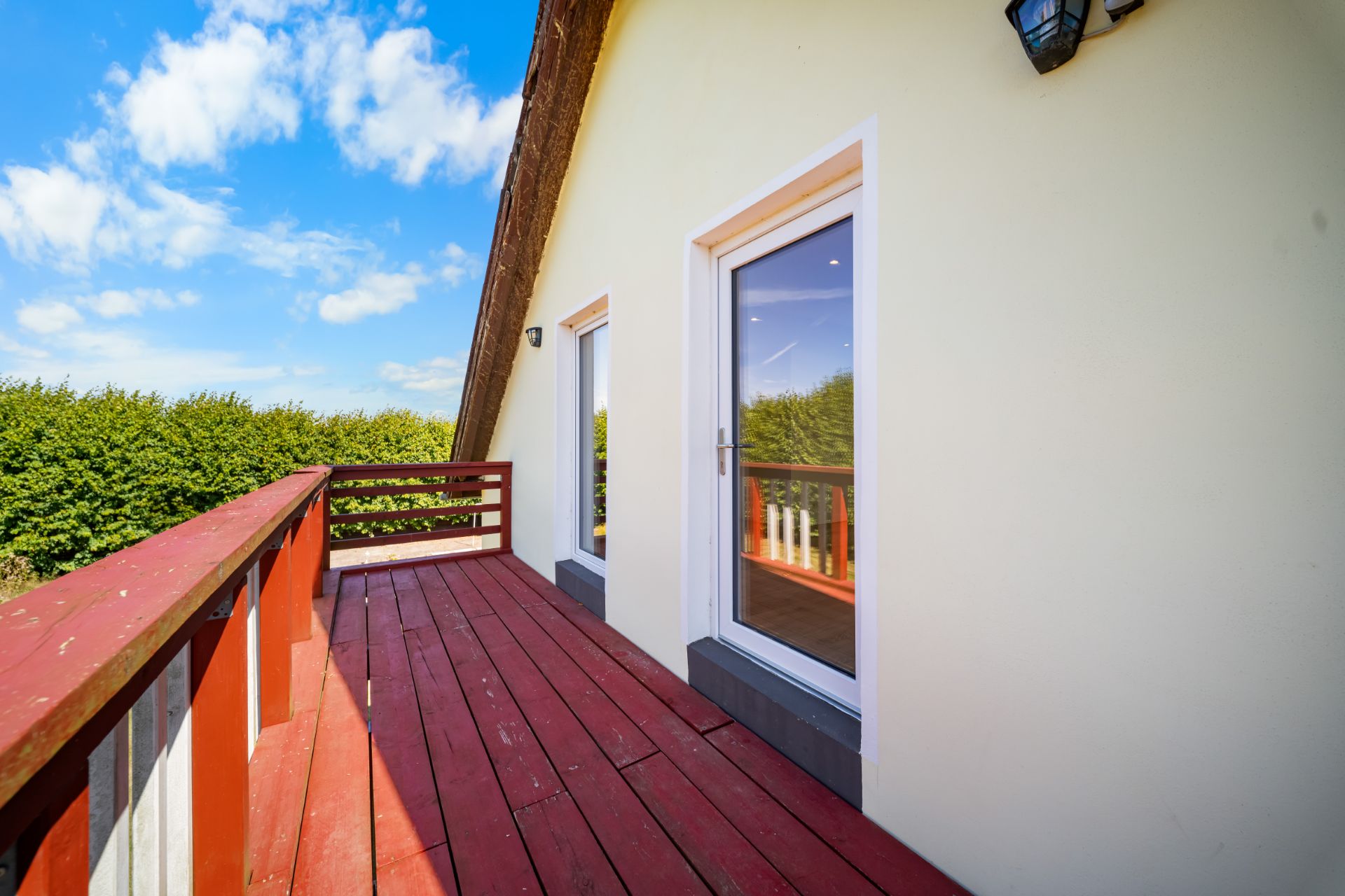 Roter Holzbalkon mit zwei Glastüren an einem hellen Haus, grüne Hecke im Hintergrund.
