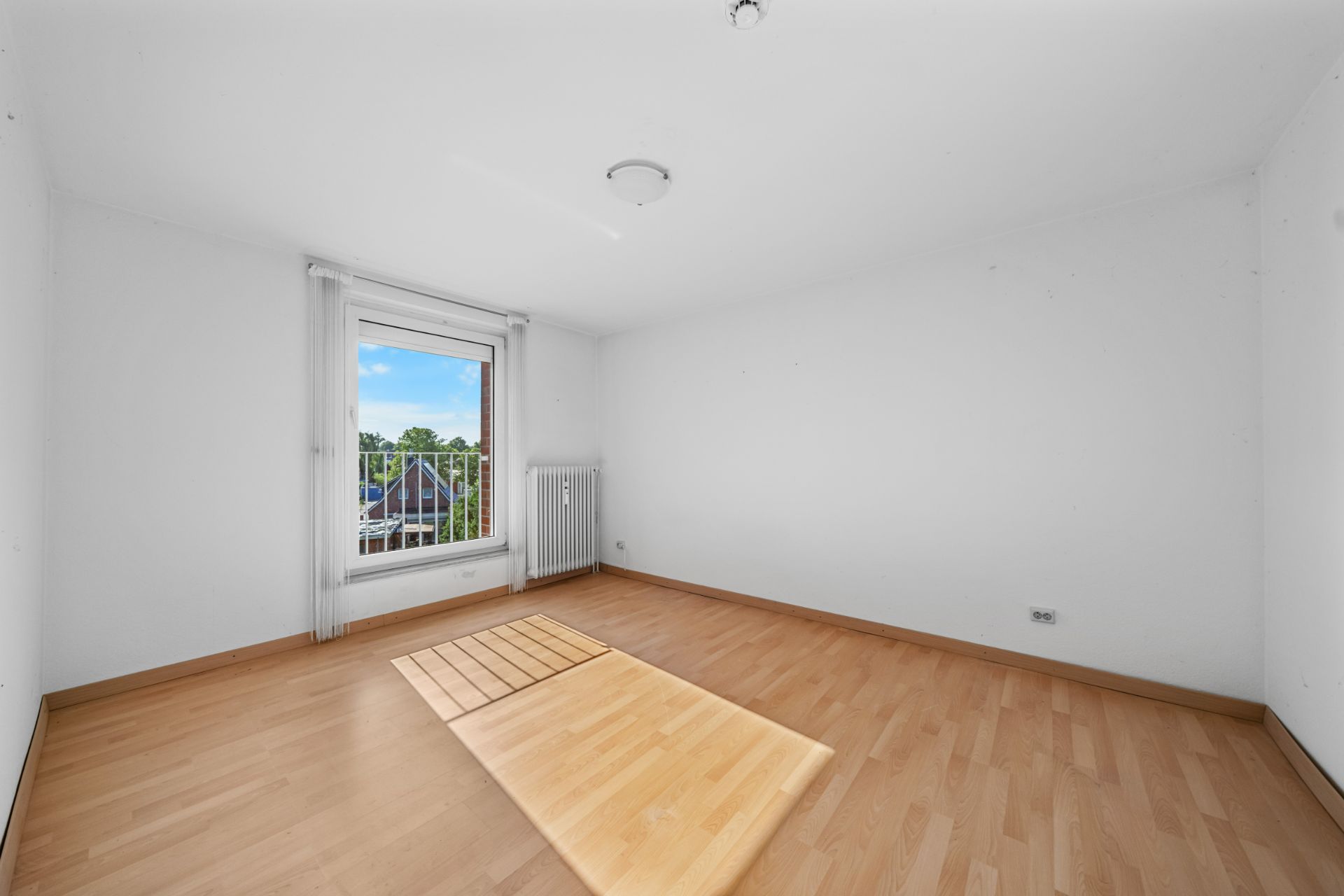 Leeres Zimmer mit Holzfußboden, großem Fenster und Heizkörper an der Wand.