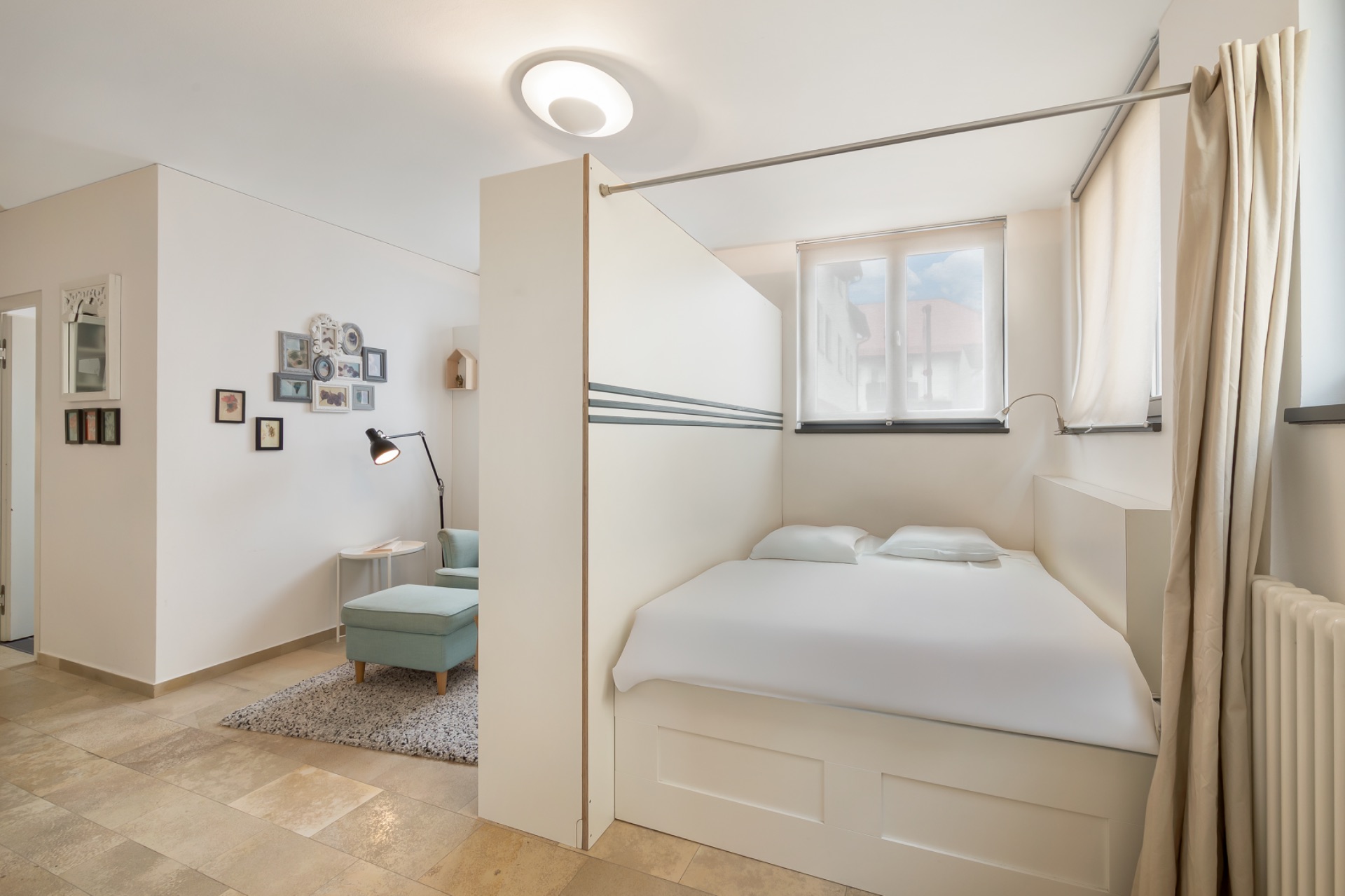 Helles Schlafzimmer mit Doppelbett, großem Fenster und Vorhang, angrenzend an einen kleinen Wohnbereich.