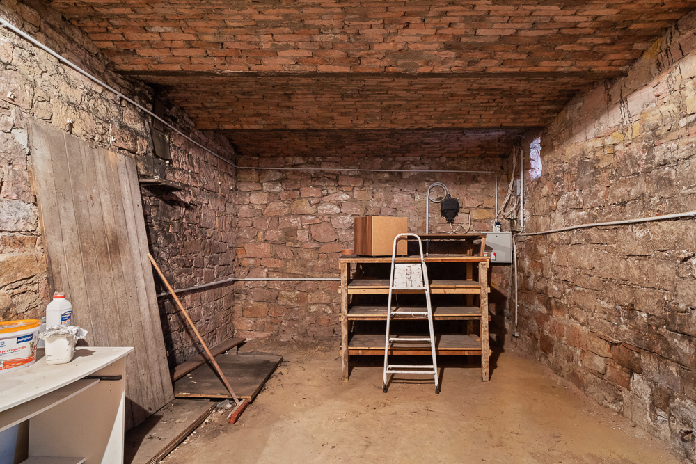 Kellerraum mit Steinwänden, Regal, Leiter und einigen Haushaltsgegenständen.