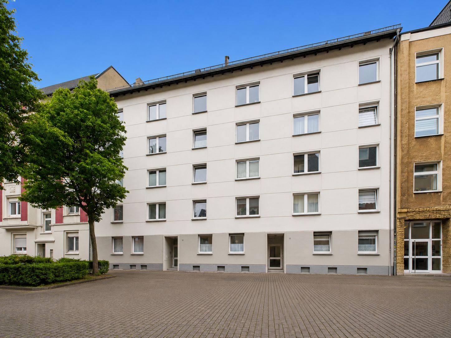Mehrstöckiges Wohnhaus mit weißer Fassade, vielen Fenstern und einem Baum davor.