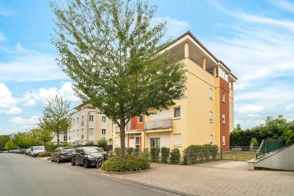 Mehrfamilienhaus mit gelber Fassade an einer ruhigen Straße, Bäume und geparkte Autos sind sichtbar.