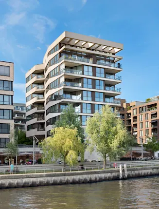 Mehrstöckige Wohngebäude mit Balkonen am Flussufer, Bäume und Gehweg im Vordergrund.