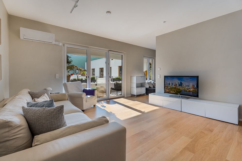Helles Wohnzimmer mit Sofa, Sessel, Fernseher und Zugang zu einer Terrasse durch große Glas-Schiebetüren.