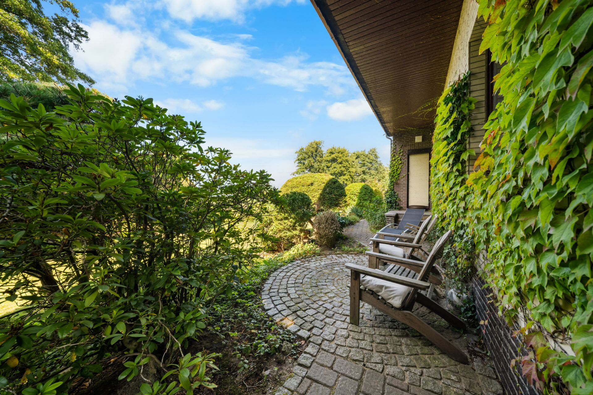 Kleine Terrasse mit zwei Stühlen, Pflastersteinen und viel grünem Bewuchs an der Hauswand.