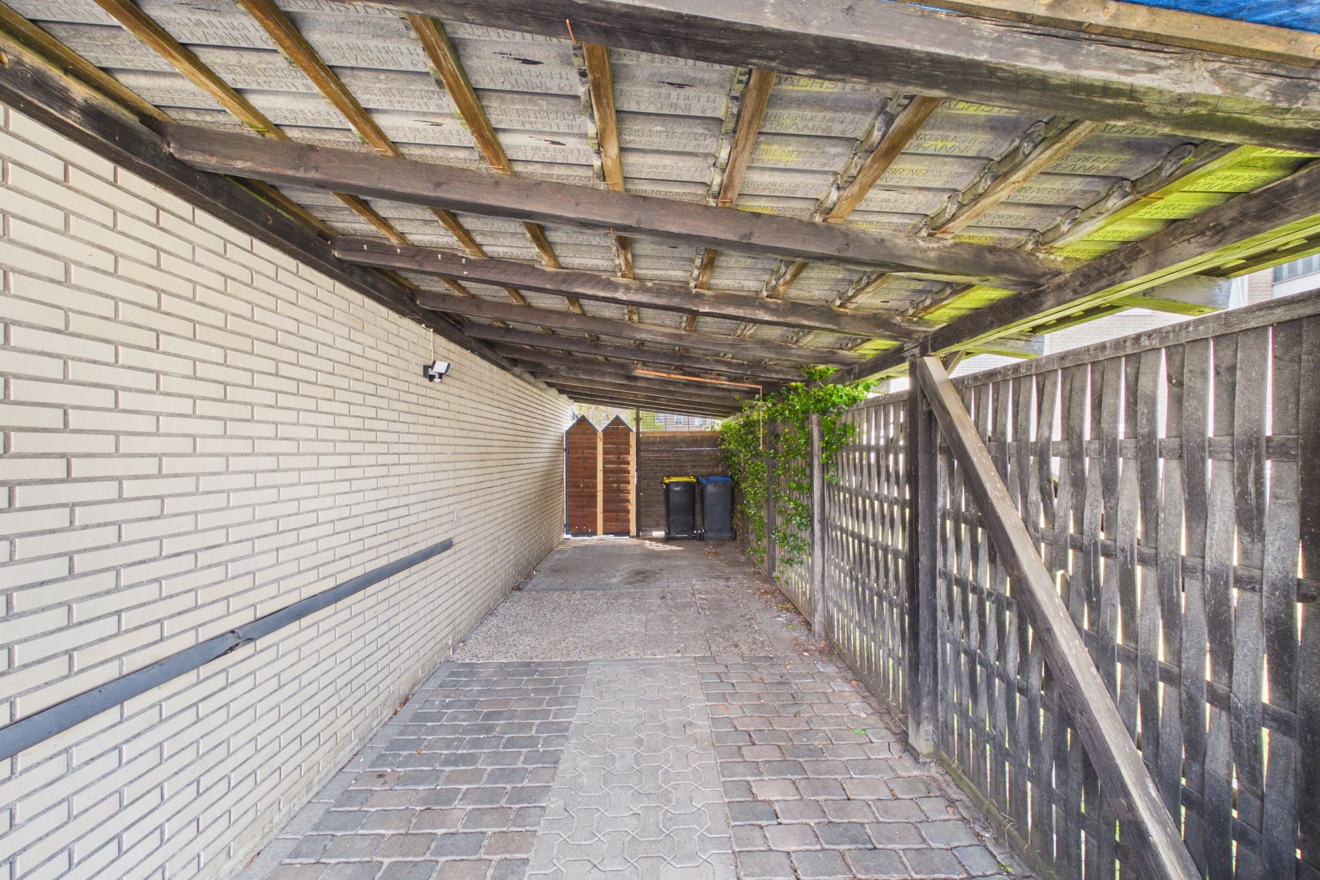 Überdachter Stellplatz mit gepflastertem Boden neben einer Hauswand und Holzzaun.