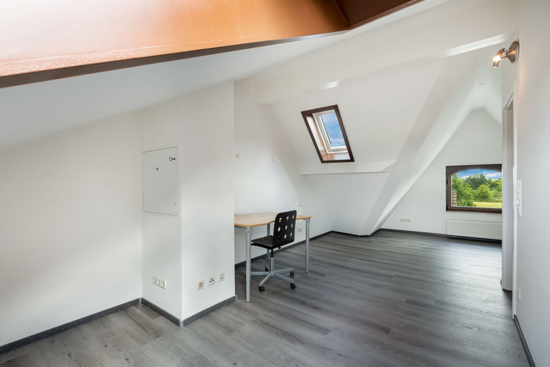 Helles Dachzimmer mit Schreibtisch, Stuhl, Dachfenster und großem Fenster mit Blick ins Grüne.