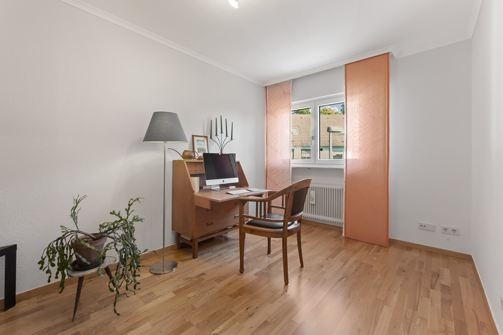 Helles Arbeitszimmer mit Holzschreibtisch, Stuhl, Computer und Stehlampe am Fenster.