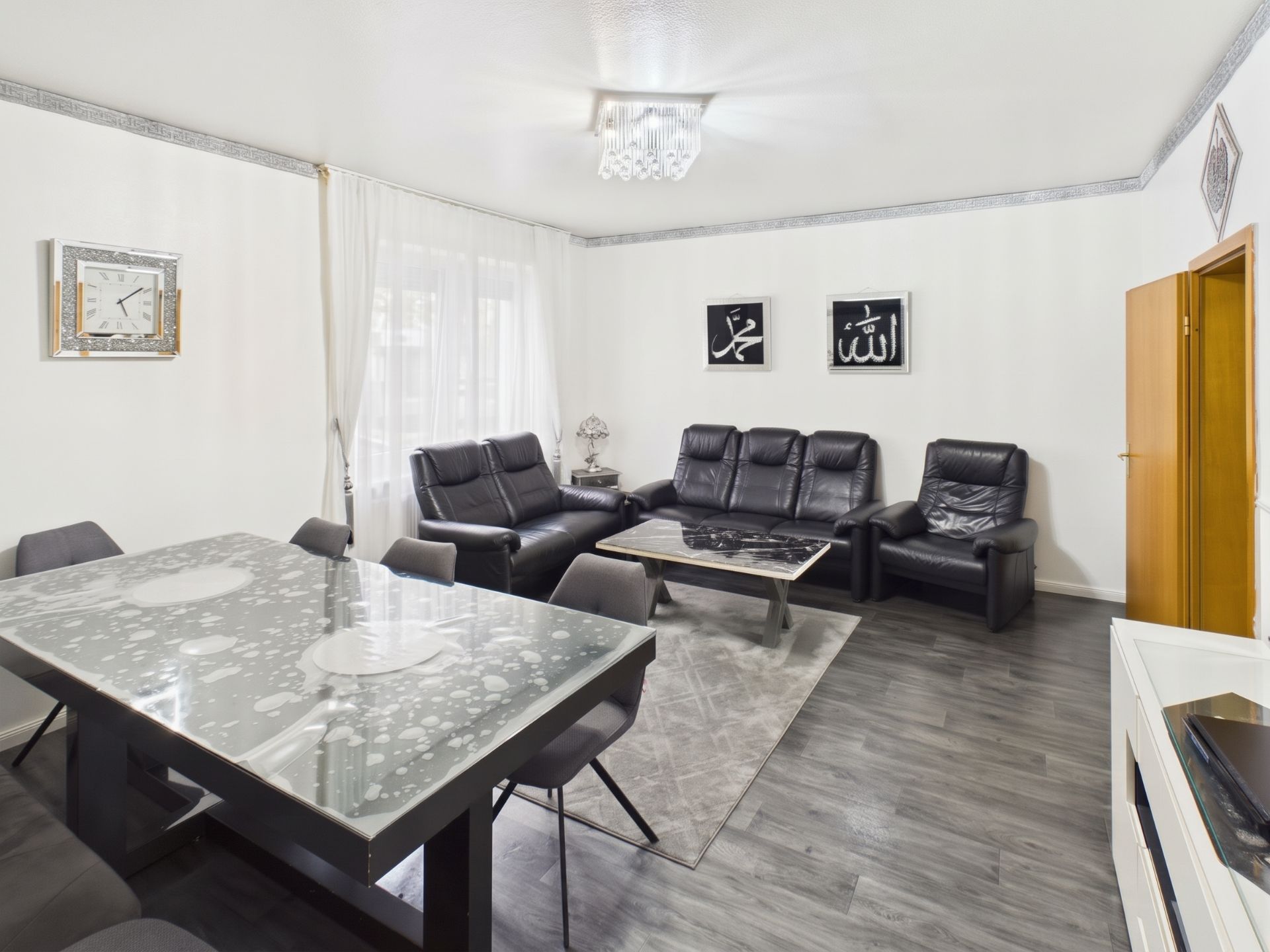 Helles Wohnzimmer mit schwarzem Ledersofa, Glastisch, Stühlen und Wandbildern.