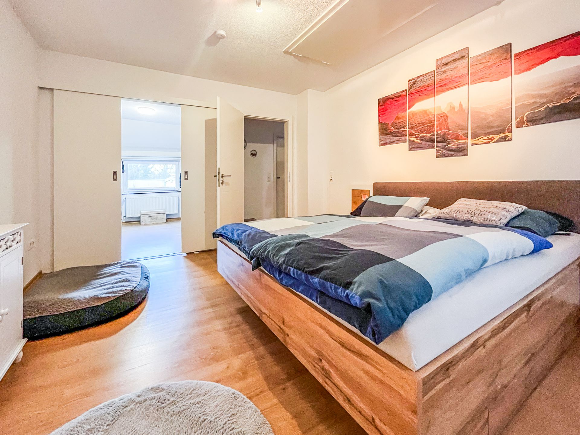 Helles Schlafzimmer mit Doppelbett, Wandbild und Holzboden, Blick in einen weiteren Raum.