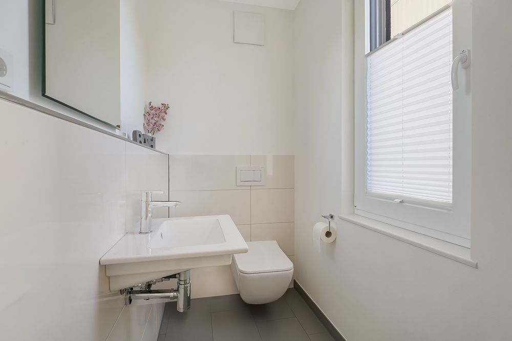 Modernes, helles Badezimmer mit Wand-WC, Waschbecken, Spiegel und Fenster mit Plissee.