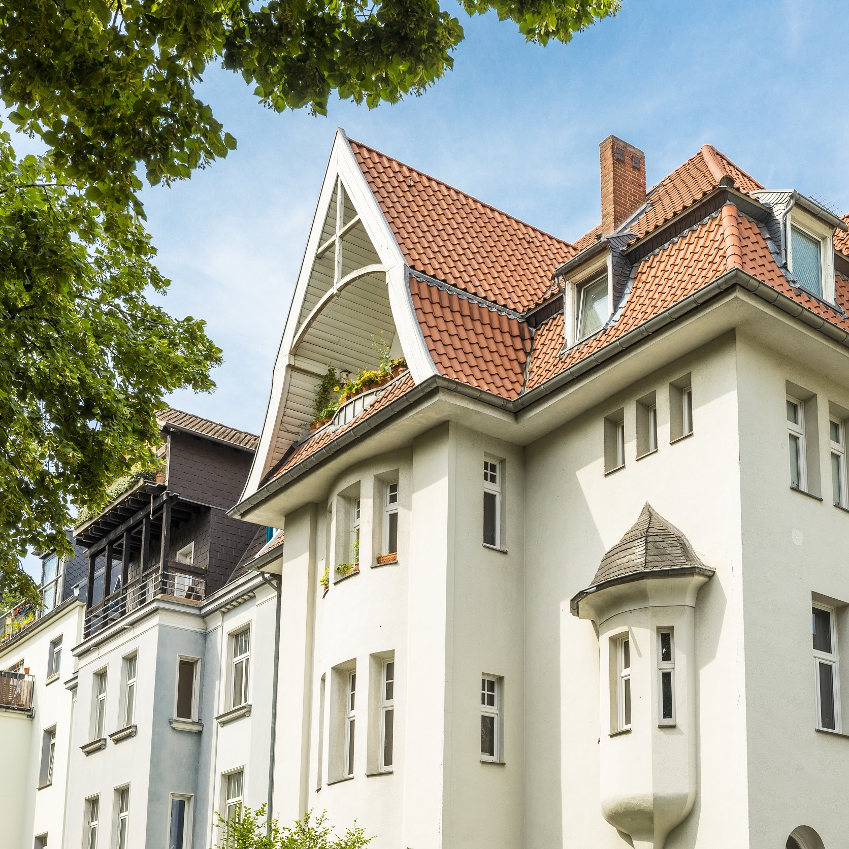 Mehrfamilienhaus mit rotem Ziegeldach und Erker, teilweise von Bäumen umrahmt.