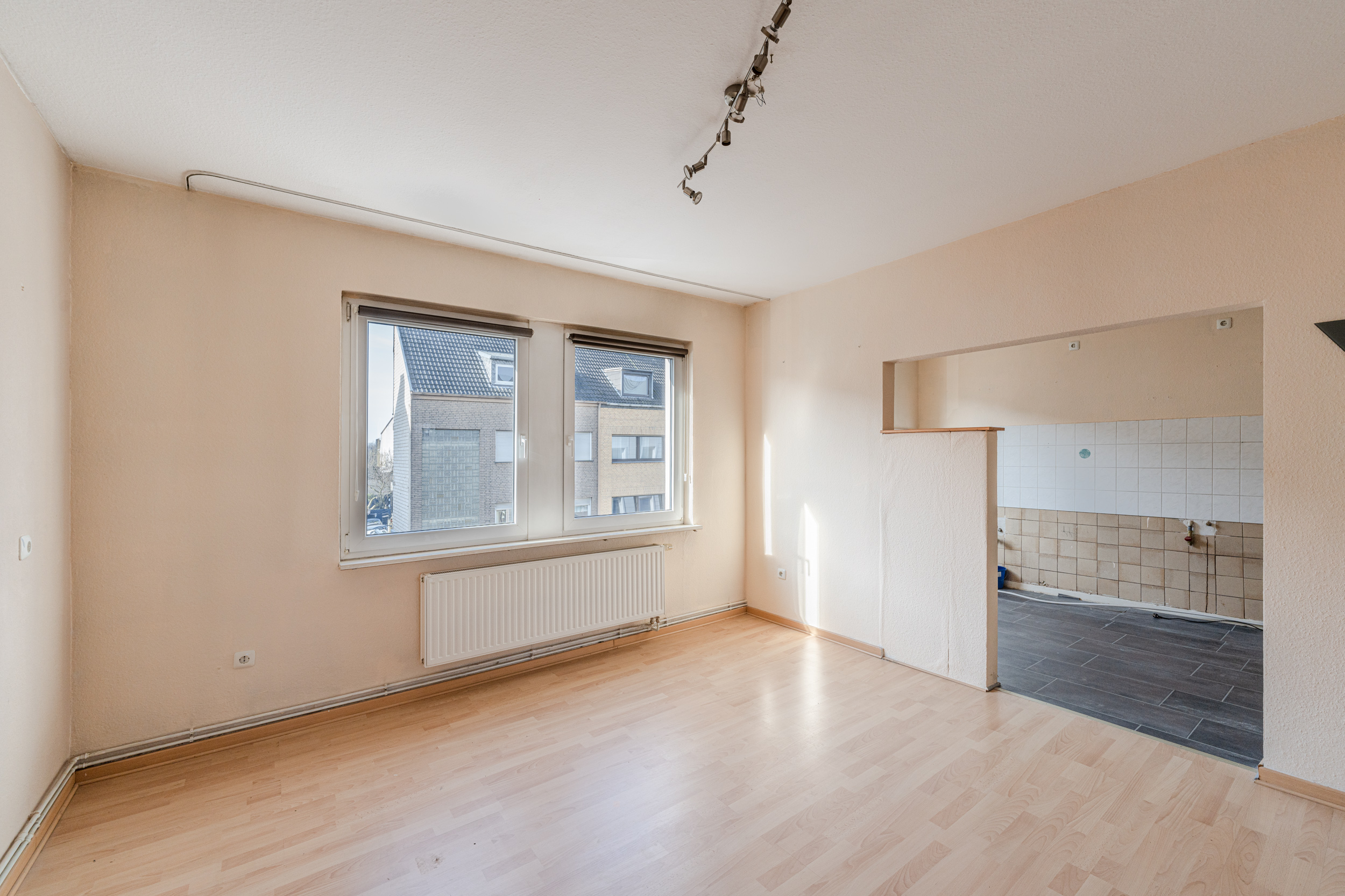Heller Raum mit zwei Fenstern, Holzfußboden und Durchgang zu gefliester Küche.
