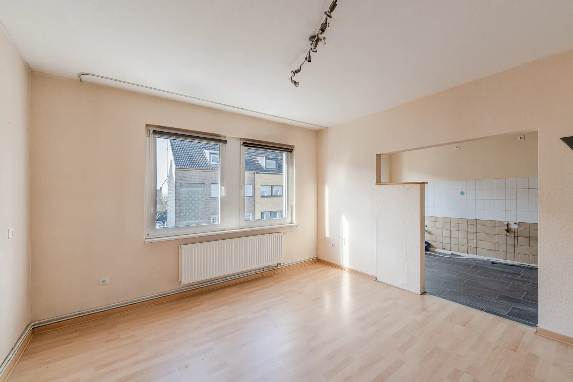 Heller Raum mit zwei Fenstern, Holzfußboden und Durchgang zu gefliester Küche.