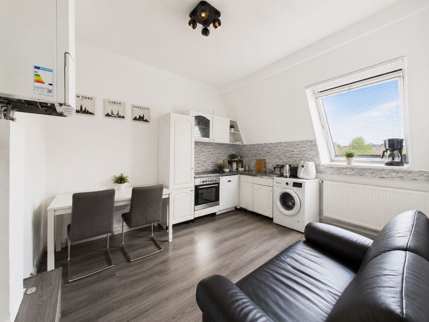 Moderne Einbauküche mit Esstisch, zwei Stühlen und schwarzem Sofa in einem hellen Raum.