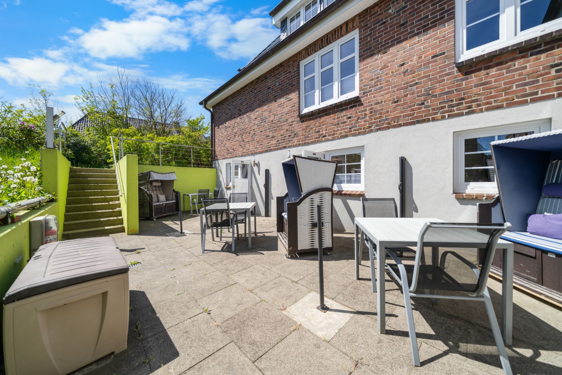 Gepflasterte Terrasse mit Gartenmöbeln und Strandkörben an einem Wohnhaus bei Sonnenschein.