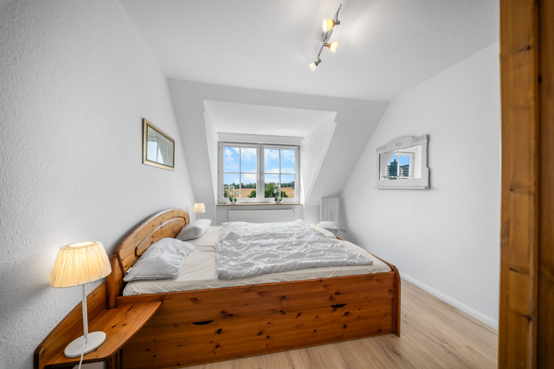 Helles Schlafzimmer mit Holzbett, Nachttisch, Lampe und großem Fenster.