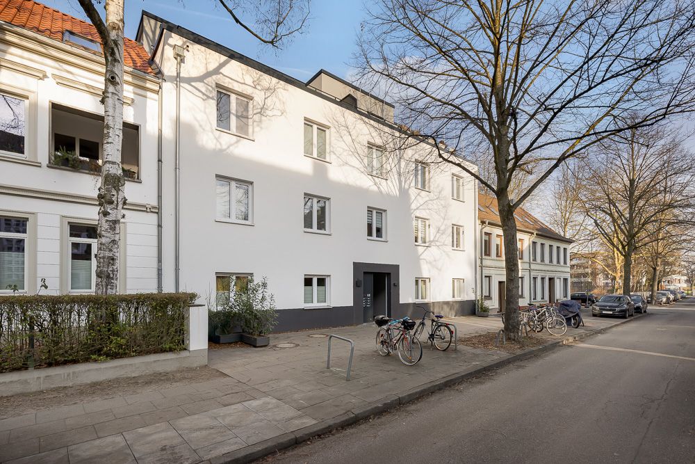 Weiße Wohnhausfassade mit mehreren Fenstern und Eingang, Fahrräder stehen auf dem Gehweg.