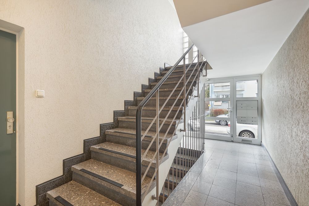 Heller Treppenhausbereich mit Steintreppe, Briefkästen und Glas-Eingangstür.