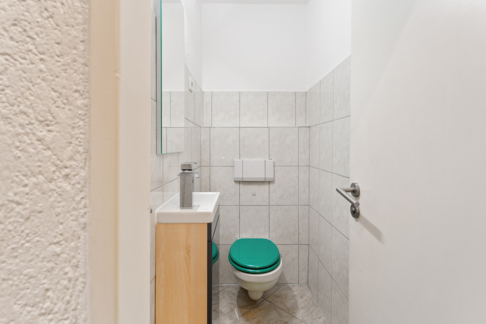 Kleines Badezimmer mit Toilette, grünem Toilettensitz und kleinem Waschbecken.