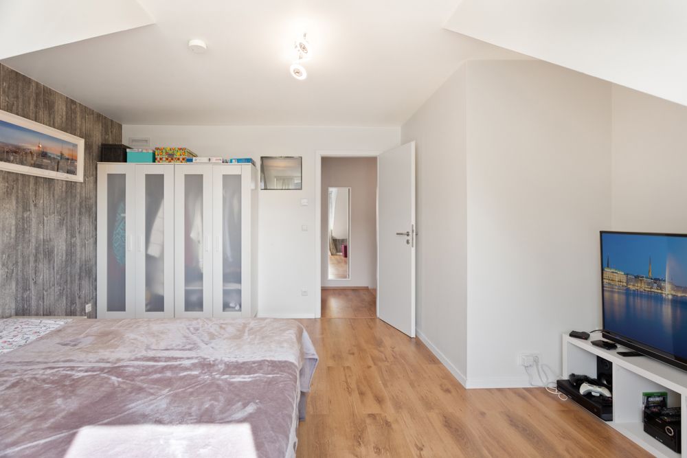 Helles Schlafzimmer mit Holzboden, Bett, Kleiderschrank und Fernseher auf weißem Regal.