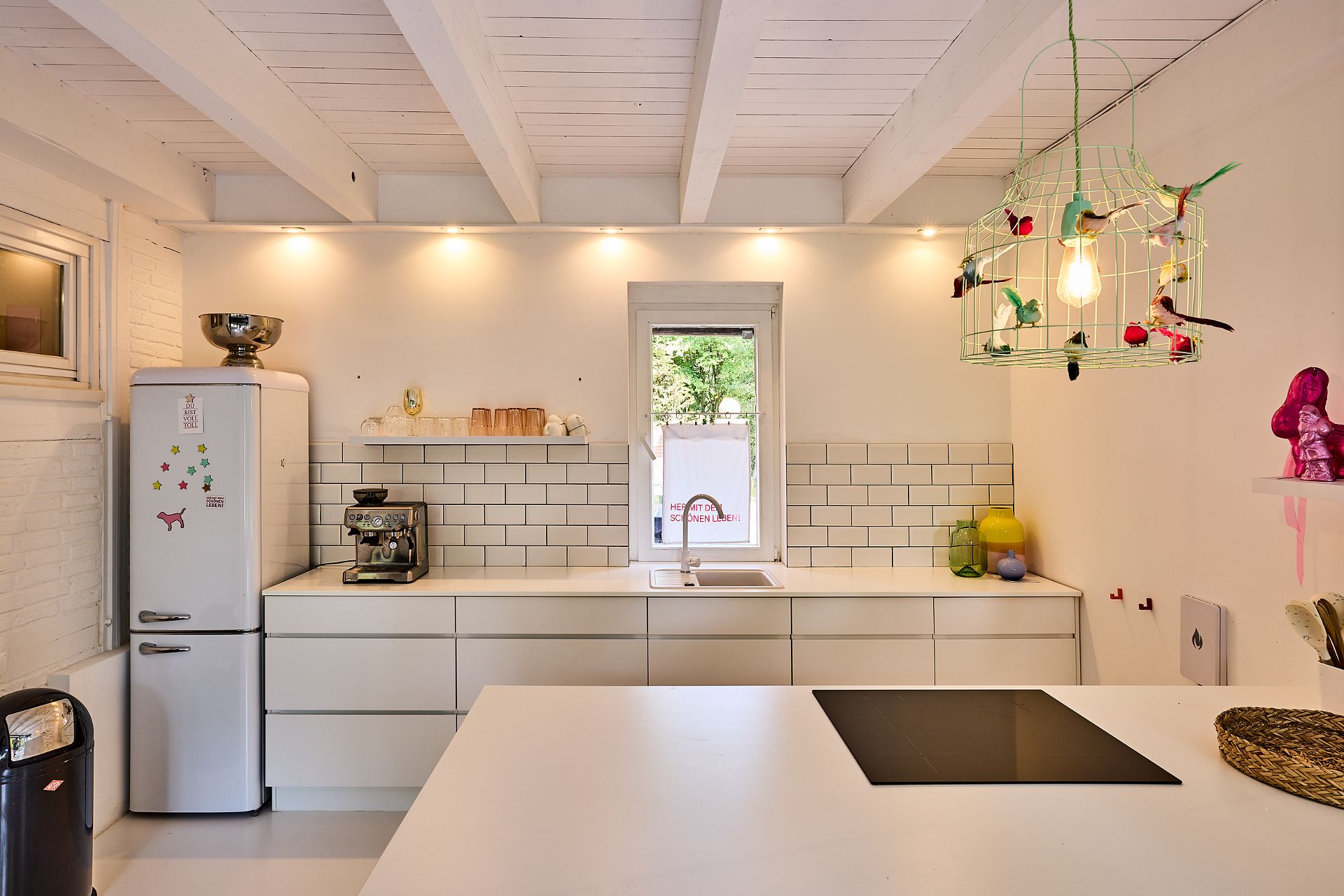 Helle Küche mit weißen Schränken, Fliesen, Fenster und bunter Lampe.