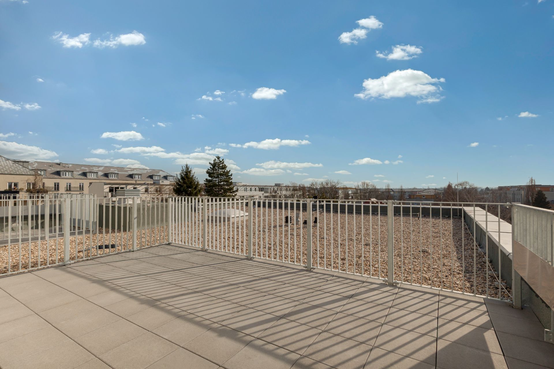 Große Terrasse mit Metallgeländer und Blick auf flaches Kiesdach und umliegende Gebäude.